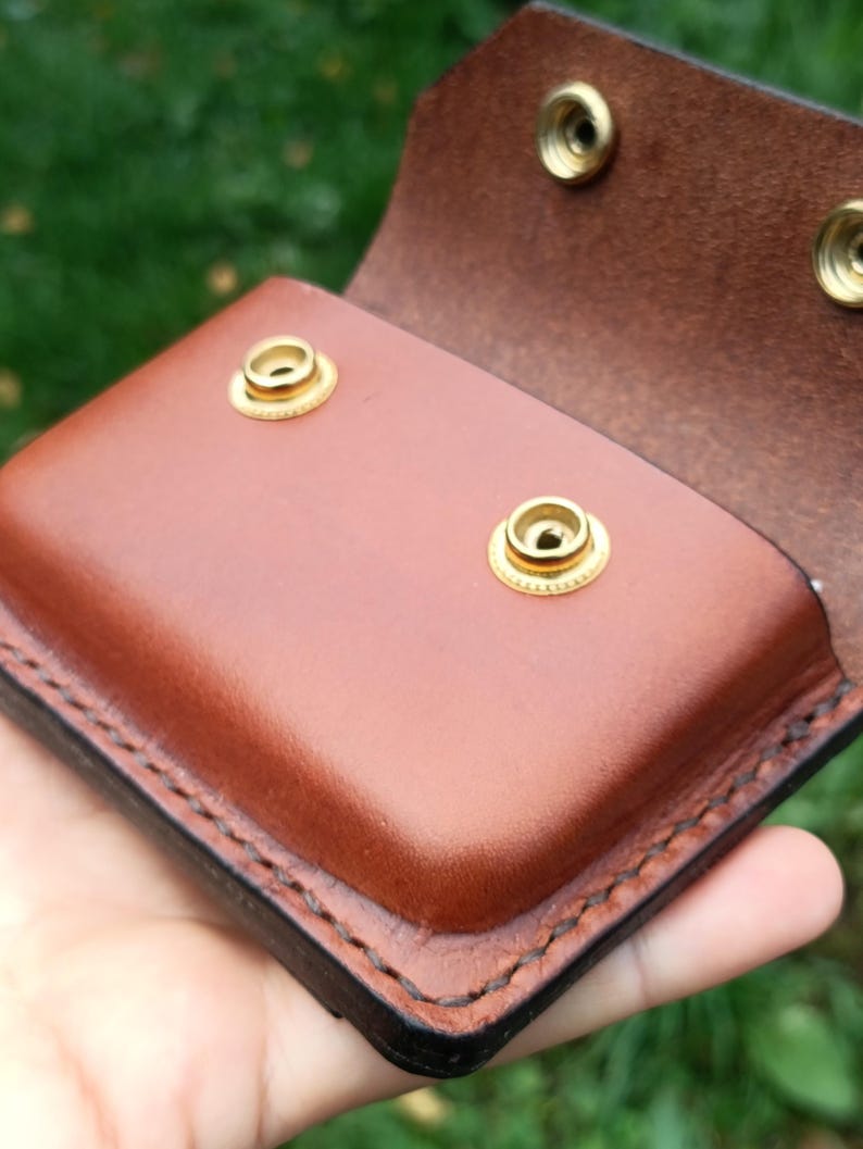Puede incluir: Una bolsa de cuero marr&oacute;n con cierre de solapa que presenta dos botones de presi&oacute;n de metal dorado en la parte frontal y dos botones a juego en la solapa interior. Los bordes est&aacute;n cosidos con hilo oscuro, mostrando un acabado liso.