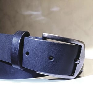 May include: Close-up of a black leather belt with a silver buckle. The belt is made of a smooth, dark material and features a classic design. The buckle is rectangular and has a brushed metal finish.