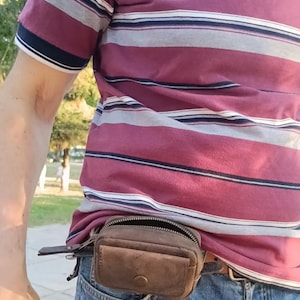 Peut inclure: Une petite pochette de ceinture en cuir marron dot&eacute;e d'un compartiment principal zipp&eacute; et d'une poche avant avec fermeture &agrave; bouton-pression. La pochette rectangulaire est fix&eacute;e &agrave; une ceinture et faite de cuir vieilli avec des coutures visibles.