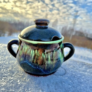 Può includere: Una piccola zuccheriera in ceramica con coperchio e due manici. La ciotola è smaltata in tonalità di verde, blu e marrone, con coperchio e manici marrone scuro. Il bordo ha un accento verde chiaro. La ciotola è appoggiata su una superficie innevata.