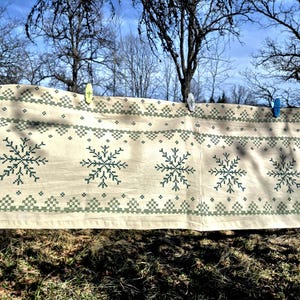 May include: A beige fabric with a repeating snowflake pattern in dark green. The fabric is held up by colorful clothespins against a backdrop of bare trees and a blue sky. The design is reminiscent of winter or holiday decor.