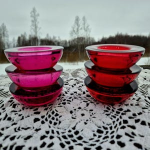 May include: Two sets of glass candle holders in the image. One set is a vibrant fuchsia, the other is a deep red. Each set has three pieces: a base and two bowl-shaped candle holders. The candle holders are sitting on a white lace doily.