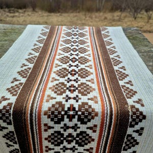 May include: A beige woven table runner with a central geometric pattern in shades of brown and orange. The runner features a repeating design of diamond and cross shapes, bordered by vertical stripes.