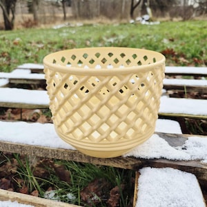 May include: A cream-colored, lattice-patterned ceramic planter. The planter has a round base and a cylindrical body with a diamond-shaped lattice design. The planter sits on a wooden surface covered in snow.