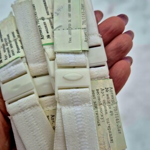 Puede incluir: Primer plano de varias correas elásticas blanquecinas con hebillas de plástico. Cada correa tiene una etiqueta con texto en un idioma extranjero. Las correas se mantienen juntas.