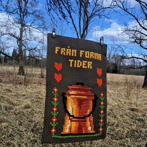 May include: A gray cross-stitch sampler with the text "Från Forna Tider" in yellow and orange lettering. The sampler features a stylized orange pot with a handle and a green floral design on either side.