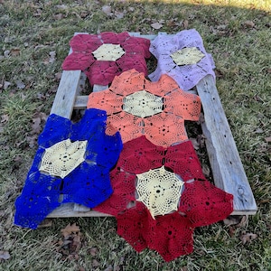 May include: A collection of colorful, handmade crochet flower doilies in shades of red, blue, orange, and purple. Each doily features a central cream-colored, open-weave design. The doilies are arranged on a rustic wooden pallet.