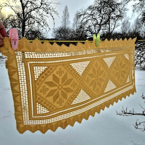 May include: A golden-yellow, rectangular lace tablecloth with a geometric pattern of diamonds and starbursts, hanging on a clothesline. The tablecloth is secured with pink and green clothespins. The background features snow and bare trees.
