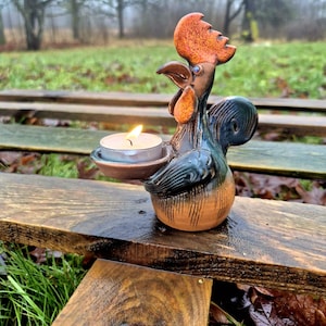 Könnte beinhalten: Ein Kerzenhalter aus Keramik in Form eines Hahns mit einer brennenden Teelichtkerze. Der Hahn ist in Orange-, Braun- und Blautönen bemalt. Der Kerzenhalter steht auf einer Holzoberfläche mit einem verschwommenen Hintergrund aus Grün.
