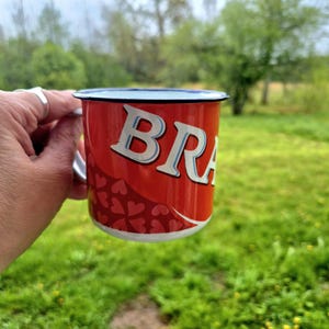 May include: A red enamel mug with the word "BRA" in white letters with a blue outline. The mug has a silver handle and a pattern of red hearts. The background is a green, grassy area.