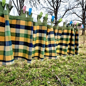 Op de afbeelding: Verschillende geruite gordijnen in geel, groen en blauw hangen aan een waslijn, vastgezet met kleurrijke wasknijpers. De gordijnen hangen buiten, met gras op de voorgrond en bomen op de achtergrond.