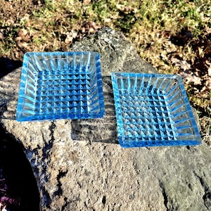 May include: Two square, light blue glass dishes with a textured, geometric pattern. The dishes are on a rough, grey stone surface, with a blurred background of grass and leaves.