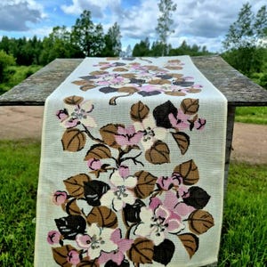 Op de afbeelding: Een rechthoekige tafelloper van gebroken witte stof met een herhalende botanische print. Het ontwerp toont gestileerde appelbloesems in tinten zachtroze en wit, geaccentueerd door donkerbruine en lichtbruine bladeren aan dunne zwarte takken.