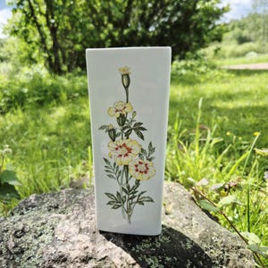 May include: A rectangular white ceramic vase with a floral design. The vase features a detailed illustration of yellow and red marigold flowers with green leaves and stems. The vase is displayed outdoors on a rock.