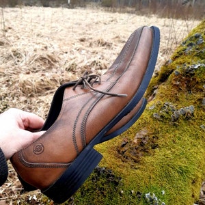 K&ouml;nnte beinhalten: Ein brauner Lederschuh mit Schn&uuml;rung und schwarzer Gummisohle. Der Schuh liegt auf einer moosbewachsenen gr&uuml;nen Oberfl&auml;che.