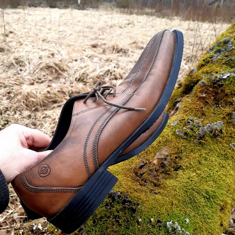 K&ouml;nnte beinhalten: Ein brauner Lederschuh mit Schn&uuml;rung und schwarzer Gummisohle. Der Schuh liegt auf einer moosbewachsenen gr&uuml;nen Oberfl&auml;che.