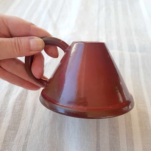 May include: A maroon ceramic mug with a unique conical shape and a rounded handle. The mug is held by a hand, showcasing its smooth, glossy finish. The design is simple and modern, suitable for coffee or tea.