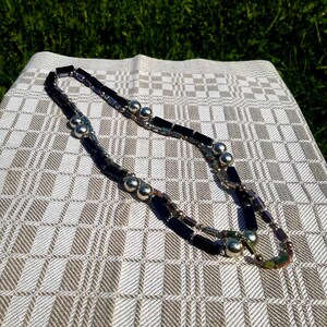 Puede incluir: Un collar de capas con cuentas rectangulares negras y cuentas esf&eacute;ricas plateadas. La joya se muestra sobre una tela a cuadros blanquecina, con hierba verde de fondo. El collar tiene un dise&ntilde;o geom&eacute;trico moderno.