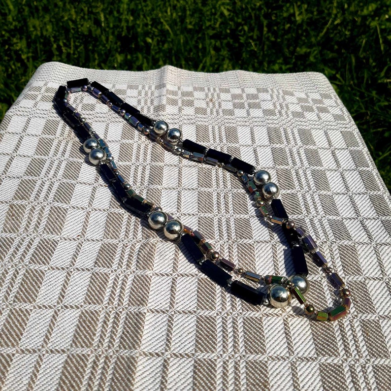 Puede incluir: Un collar de capas con cuentas rectangulares negras y cuentas esf&eacute;ricas plateadas. La joya se muestra sobre una tela a cuadros blanquecina, con hierba verde de fondo. El collar tiene un dise&ntilde;o geom&eacute;trico moderno.