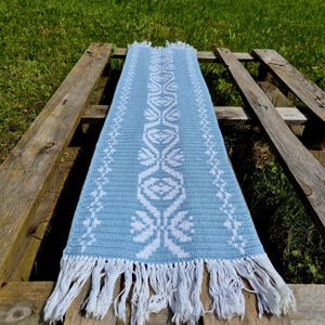 Può includere: Un runner da tavolo tessuto azzurro con un motivo floreale bianco e frange. Il runner rettangolare è appoggiato su un pallet di legno all'aperto. Le frange sono bianche.