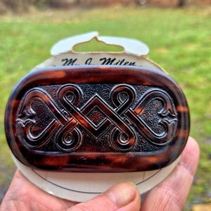 Puede incluir: Pinza de pelo ovalada con estampado de carey y un intrincado diseño de nudo celta marrón oscuro. La pinza tiene un fondo oscuro texturizado y está adherida a una tarjeta blanca con el texto "M. A. Miles".