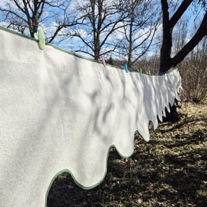 May include: A white, scalloped-edge textile, possibly a rug or blanket, hangs on a clothesline outdoors. The edge is trimmed with green. Several colorful clothespins secure the textile to the line. The background features trees and a blue sky.