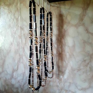 Puede incluir: Varias collares cuelgan de ganchos. Los collares presentan una combinaci&oacute;n de cuentas rectangulares negras, cuentas esf&eacute;ricas plateadas y cuentas iridiscentes m&aacute;s peque&ntilde;as. Se exhiben contra una pared veteada.