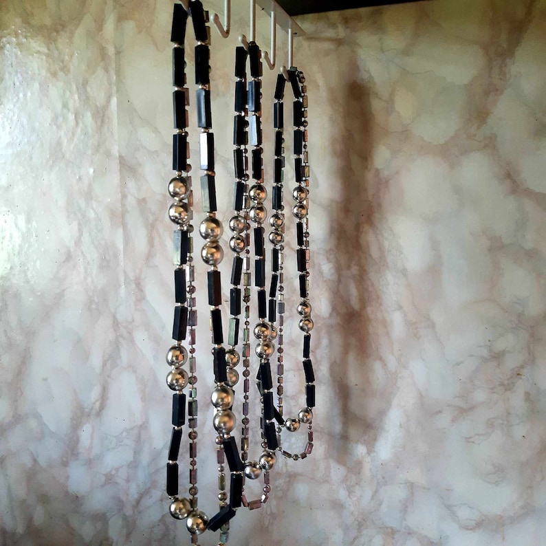 Puede incluir: Varias collares cuelgan de ganchos. Los collares presentan una combinaci&oacute;n de cuentas rectangulares negras, cuentas esf&eacute;ricas plateadas y cuentas iridiscentes m&aacute;s peque&ntilde;as. Se exhiben contra una pared veteada.