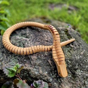 Pode incluir: Um brinquedo de cobra de madeira artesanal, esculpido em madeira de cor clara. A cobra é segmentada, permitindo um movimento flexível, e está pousada em uma rocha cinza com um fundo verde desfocado. A cabeça da cobra é detalhada.