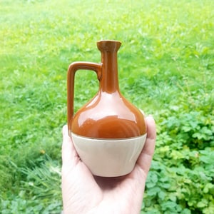 Può includere: Un piccolo boccale in ceramica marrone e bianca con un manico. Il boccale ha un corpo rotondo e un collo stretto.