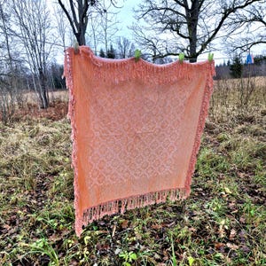 Puede incluir: Una manta tejida de color melocotón con flecos cuelga de un tendedero. La manta tiene un patrón texturizado y está asegurada con pinzas verdes. El fondo presenta árboles y follaje, lo que sugiere un entorno al aire libre.