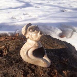 Puede incluir: Figura de cerámica beige claro de un mono con sombrero, con la boca abierta y expresión de sorpresa. La figura tiene una gran abertura en la parte delantera. El mono está colocado sobre una roca. Objeto decorativo.