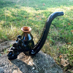 May include: A black ceramic pipe with a decorative dragon design and a brown glazed vase on top. The pipe is sitting on a rock.