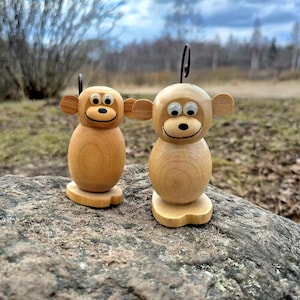 以下が含まれることがあります： 目は塗られていて、笑顔の顔をした木製の猿の置物2つ。猿は岩の上に立っています。
