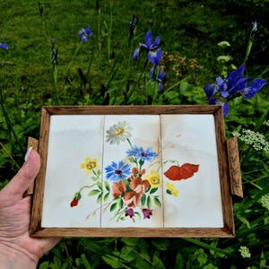 Può includere: Un vassoio rettangolare con cornice e manici in legno. La superficie del vassoio è divisa in tre sezioni, ciascuna con un motivo floreale ad acquerello con fiori blu, gialli, rossi e marroni. Lo sfondo è bianco.