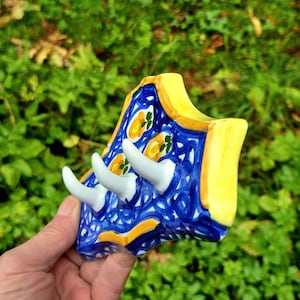 May include: A ceramic wall hook rack featuring a blue and white swirling pattern with yellow floral accents and a solid yellow border. The piece includes three white, curved, horn-shaped hooks extending from the front of the decorative base.
