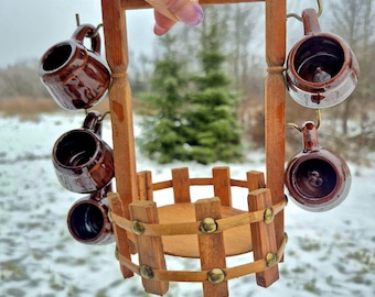 May include: A miniature wooden well-shaped mug holder with six small brown ceramic mugs hanging from hooks. The well has a round base and a fence-like structure. The background is a snowy outdoor scene.