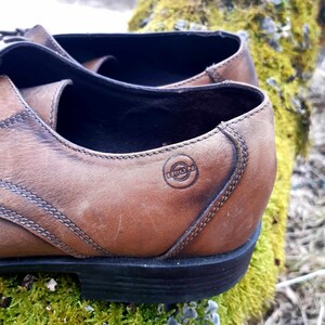 K&ouml;nnte beinhalten: Nahaufnahme eines braunen Lederschuhs mit schwarzer Sohle. Der Schuh hat ein gen&auml;htes Design und ein kreisf&ouml;rmiges Logo mit dem Text "KOD" darin.