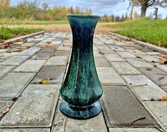May include: A ceramic vase with a flared rim and a wide base, featuring a mottled glaze in shades of deep forest green, emerald, and dark navy blue. The surface has a glossy finish with vertical streaks and a smooth, curved silhouette.