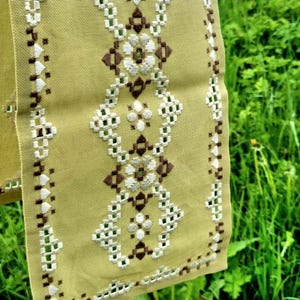 May include: Close-up of a section of embroidered fabric. The fabric is a pale yellow-green color, with a repeating pattern of white, brown, and green floral and geometric designs. The embroidery is detailed and intricate.