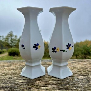 Può includere: Una coppia di vasi in porcellana bianca con bordo svasato e base quadrata. Ogni vaso presenta un motivo dipinto a mano di piccoli fiori di viola del pensiero viola e gialli con steli verdi sparsi sulla superficie bianca liscia e lucida.