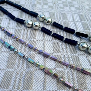 Puede incluir: Tres collares con diferentes dise&ntilde;os. El collar superior tiene cuentas rectangulares negras y cuentas esf&eacute;ricas plateadas. Los dos collares inferiores tienen cuentas rectangulares iridiscentes y cuentas esf&eacute;ricas plateadas. Los collares est&aacute;n sobre una superficie beige estampada.