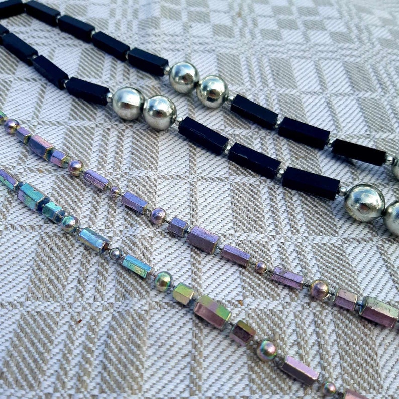Puede incluir: Tres collares con diferentes dise&ntilde;os. El collar superior tiene cuentas rectangulares negras y cuentas esf&eacute;ricas plateadas. Los dos collares inferiores tienen cuentas rectangulares iridiscentes y cuentas esf&eacute;ricas plateadas. Los collares est&aacute;n sobre una superficie beige estampada.