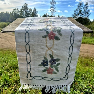 May include: A rectangular white fabric table runner featuring a central embroidered floral pattern in shades of pink, blue, and green, framed by a dark blue chain-link border design, finished with a fringed edge on the bottom end.