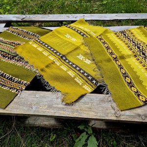 May include: Three vintage woven textile pieces in shades of green and yellow. Each piece features intricate geometric patterns in white, black, and brown. The textiles have a fringed edge and are arranged on a weathered wooden surface.