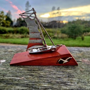 Puede incluir: Una escultura de velero en miniatura con una base de madera roja. El barco es plateado con una vela roja y blanca. Una gaviota blanca está tallada en la base. La escultura mide aproximadamente 13 cm de largo.
