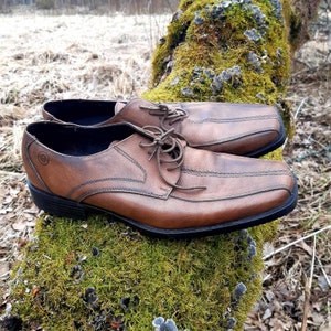 K&ouml;nnte beinhalten: Ein Paar braune Lederschuhe mit Schn&uuml;rung und einem gen&auml;hten Design. Die Schuhe liegen auf einem Bett aus gr&uuml;nem Moos.