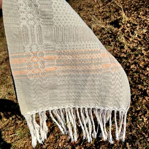 Könnte beinhalten: Gewebter Textil mit einem hellgrauen und pfirsichfarbenen geometrischen Muster. Der Textil hat einen Fransen am unteren Rand. Der Textil ist über einer Oberfläche mit braunen Blättern drapiert.