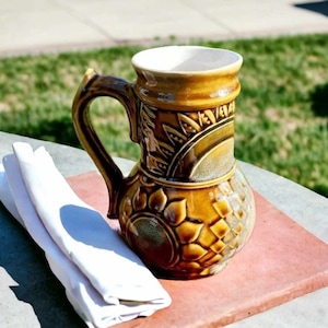 Può includere: Un'anfora in ceramica marrone con un motivo floreale. L'anfora ha un manico e una bocca larga. È appoggiata su una superficie di piastrelle marroni.