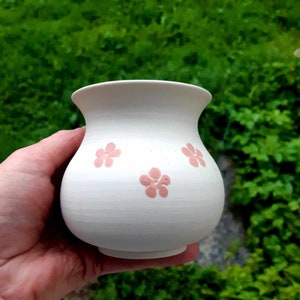 Può includere: Vaso in ceramica bianca con corpo arrotondato e un'ampia apertura. Il vaso presenta tre motivi di fiori rosa sul davanti.